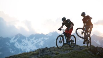 Zwei Mountainbiker stehen auf einem Hügel und genießen die Aussicht auf die schneebedeckten Berge. Der Himmel ist bewölkt und die Szene wirkt abenteuerlich.