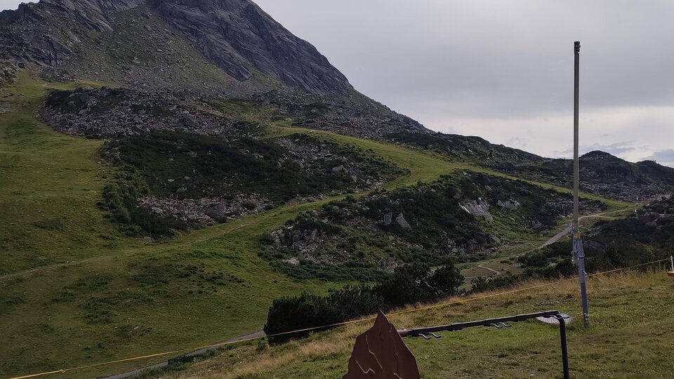 A green mountain landscape with a high peak in the background. In the foreground, there is a sign or information board.