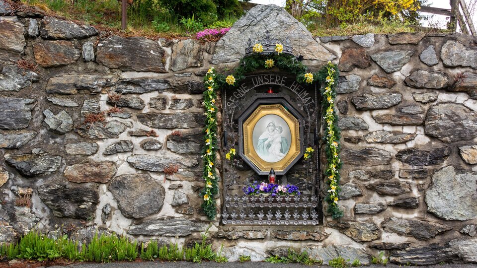 A stone wall with a decorated portrait in a niche. The image is adorned with flowers and depicts a religious scene. | © Bernhard Gruber – Photography