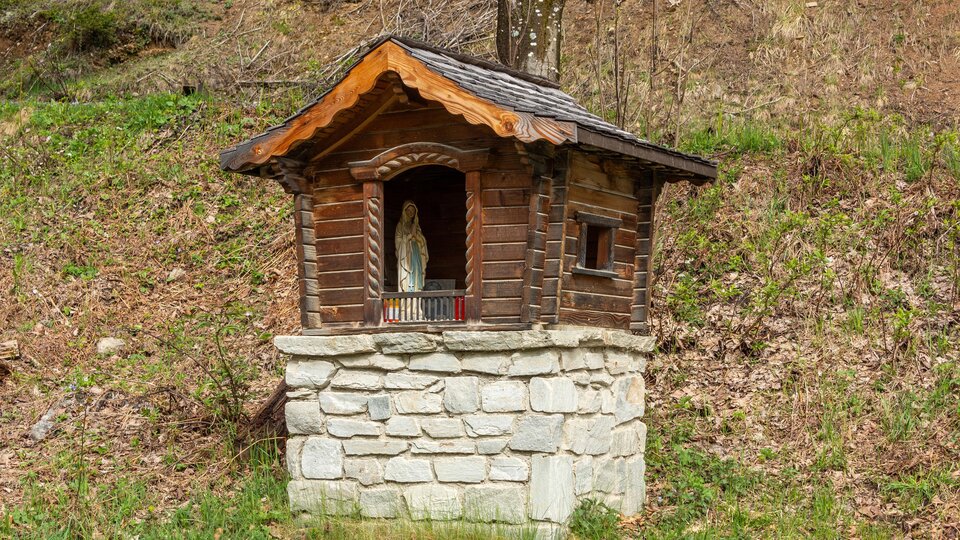 A small wooden chapel stands on a stone pedestal in nature. Next to the chapel, green meadows and foliage can be seen. | © Bernhard Gruber – Photography