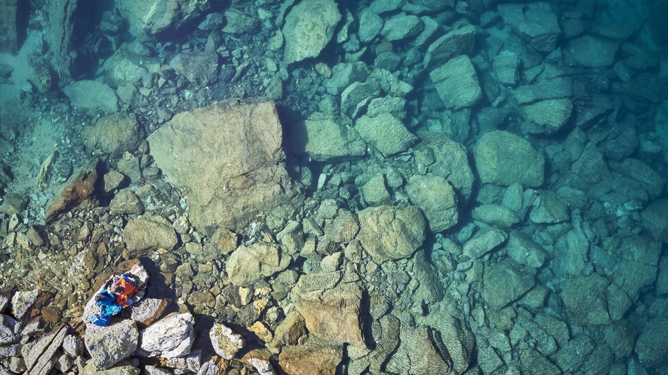 A clear blue lake with many large stones at the bottom. A person is lying relaxed on the shore.