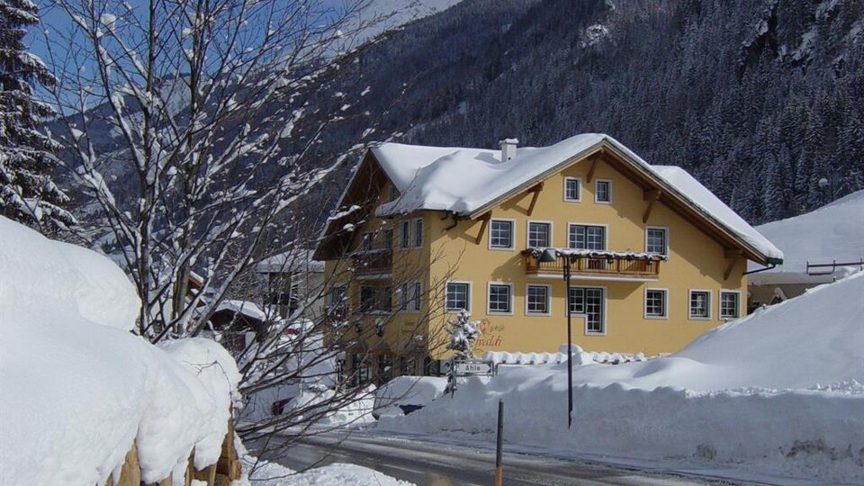 A charming yellow house in the snow with a picturesque mountain backdrop. The path is covered in fresh snow and the trees are decorated for winter.