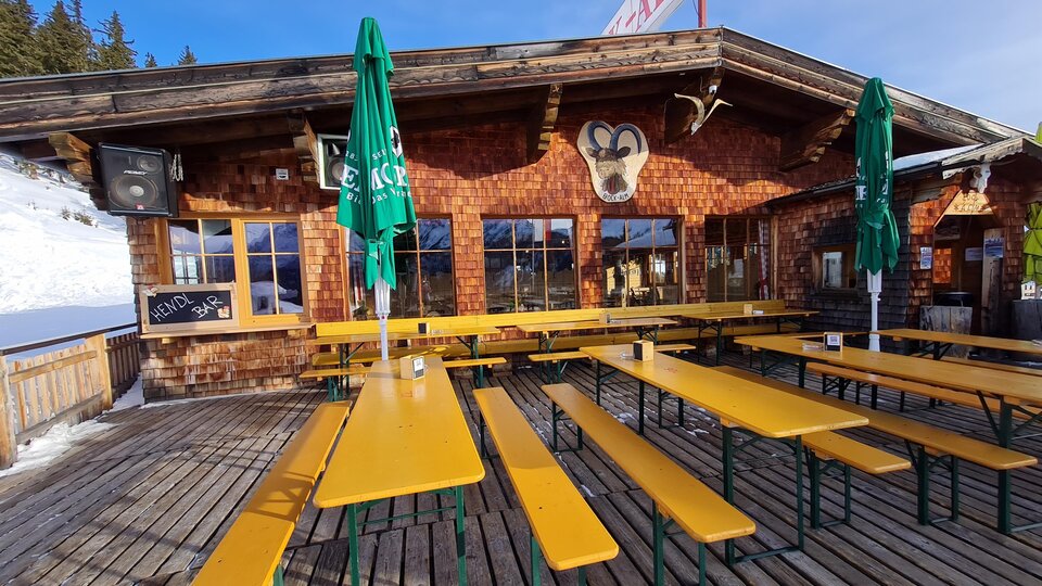 A cozy mountain cabin with wood cladding and large windows. Outside, there are long benches and tables with green umbrellas.