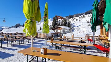 A cozy terrace with wooden tables and colorful parasols in the snow. In the background, there are snow-covered mountains and a cable car visible.
