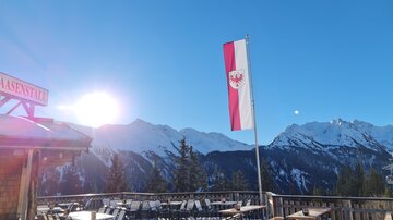 A beautiful view of snow-covered mountains under a clear blue sky. In the foreground, there are tables and a flag is waving in the wind.