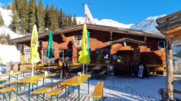 A mountain cabin with a wooden facade and several tables outdoors. In the background, there are snow-covered mountains and a blue sky.