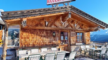 A cozy mountain cabin with wooden cladding and outdoor tables. The sky is clear and blue, promising a beautiful view of the mountains.