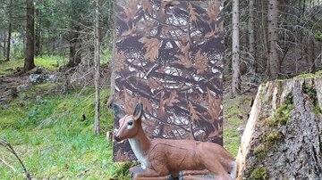 A deer made of plastic lies in front of a camouflage-colored wall in a forest. The surroundings consist of green plants and trees.