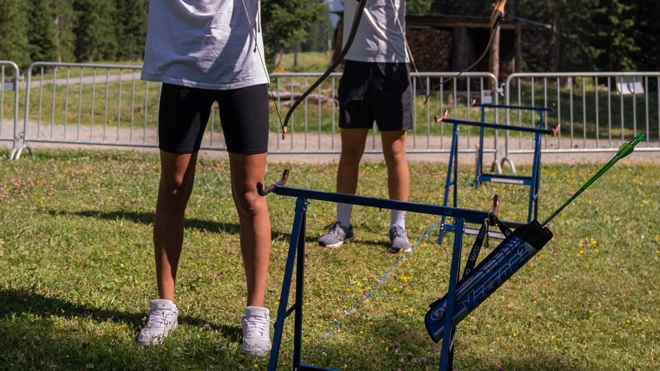Two teenagers are practicing archery in a beautiful, natural setting. Mountains and trees are visible in the background.