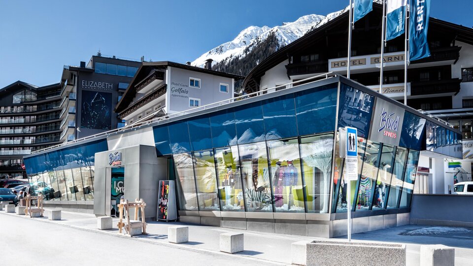 A modern building with large glass windows in an Alpine landscape. In the background, snow-covered mountains and typical alpine architecture can be seen.
