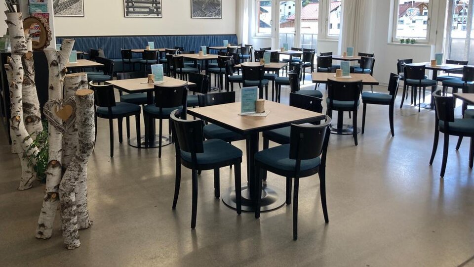 A modern restaurant with tables and chairs. The walls are bright and large windows let in plenty of light.