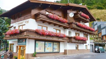 A charming house with blooming flower boxes and an inviting café door. In the background, green hills can be seen.
