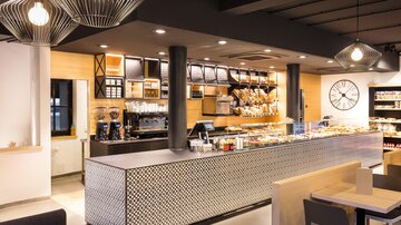 Ein modernes Café mit einem auffälligen Tresen und einer einladenden Atmosphäre. Die Einrichtung ist stilvoll und hell, mit verschiedenen Verkaufsregalen.