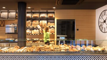 Eine moderne Bäckerei mit einer Auswahl an frischem Brot und Gebäck. Im Hintergrund ist eine Mitarbeiterin zu sehen, die die Regale auffüllt.