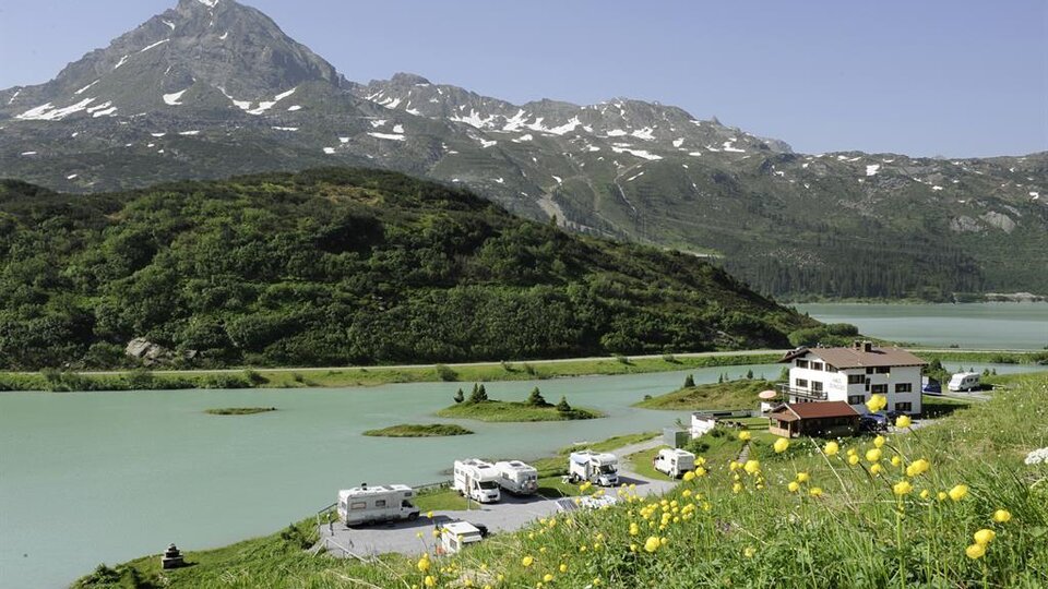 A picturesque landscape with a calm lake and an impressive mountain in the background. At the shore, RVs and a guesthouse are surrounded by green meadows.