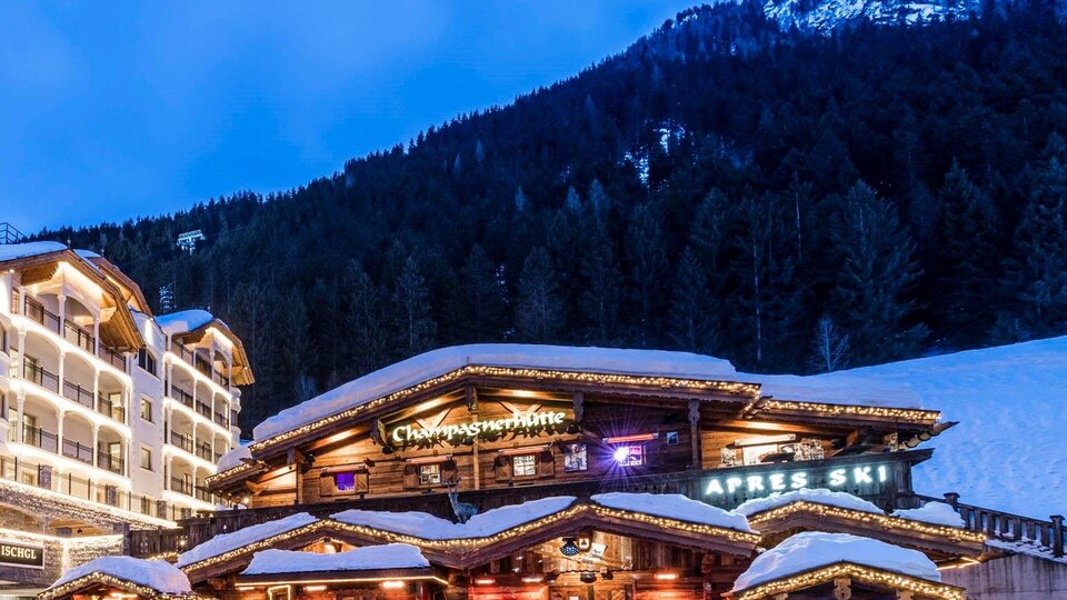 A cozy mountain cabin in the snow with lights that shine in the twilight. In the background, there are snow-covered mountains and forests.