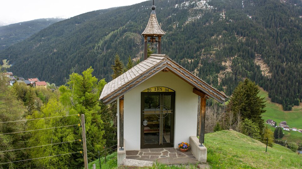 A small chapel stands on a hill, surrounded by trees and mountains. In the background, a picturesque mountain landscape can be seen. | © Bernhard Gruber – Photography