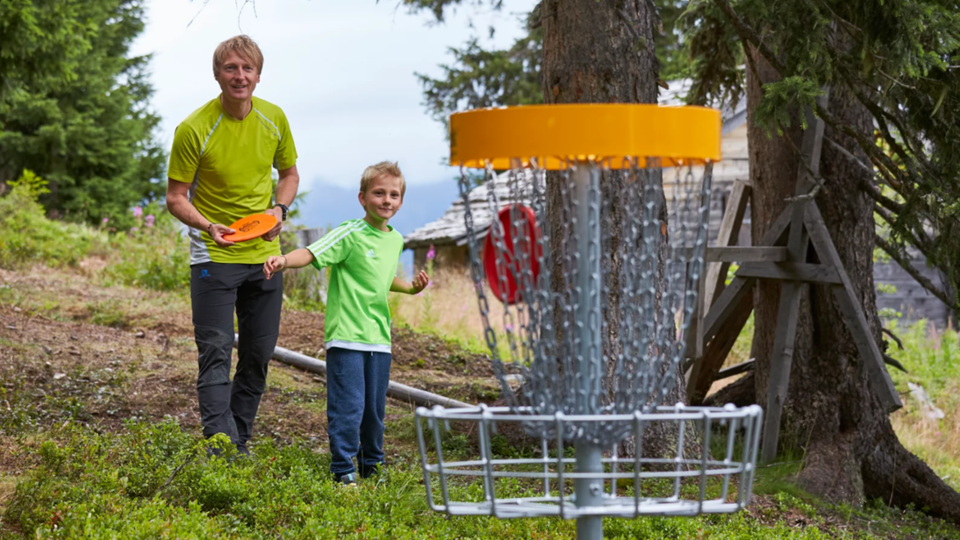 A man and a boy are standing in a forest and playing disc golf. In the background, the basket for the game can be seen.