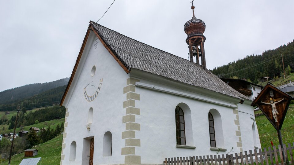 A small, white church with a tall tower stands in a green landscape. In the background, gentle hills and forests can be seen. | © Bernhard Gruber – Photography