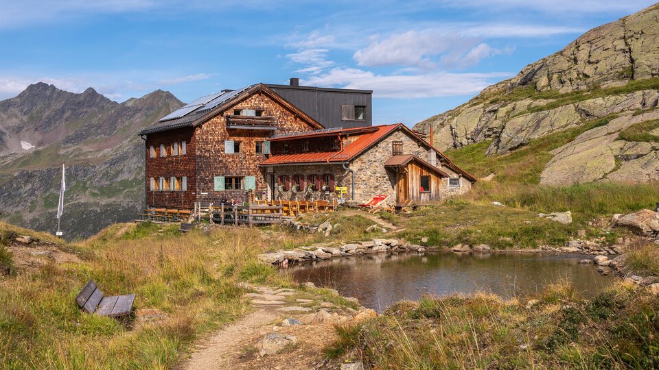 A picturesque mountain cabin in the midst of a green landscape. In the foreground, there is a small pond, and in the background, impressive mountains can be seen.