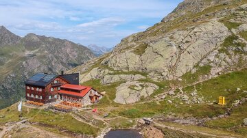 A mountain cabin is picturesque set in a mountainous landscape. In the background, there are tall mountains and a clear sky.