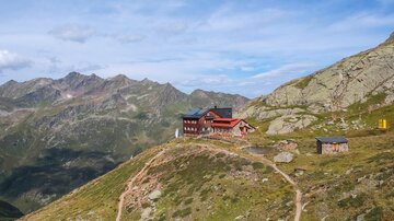 An impressive mountain landscape with a mountain hut on a summit. The sky is partly cloudy and the surrounding mountains are clearly visible.