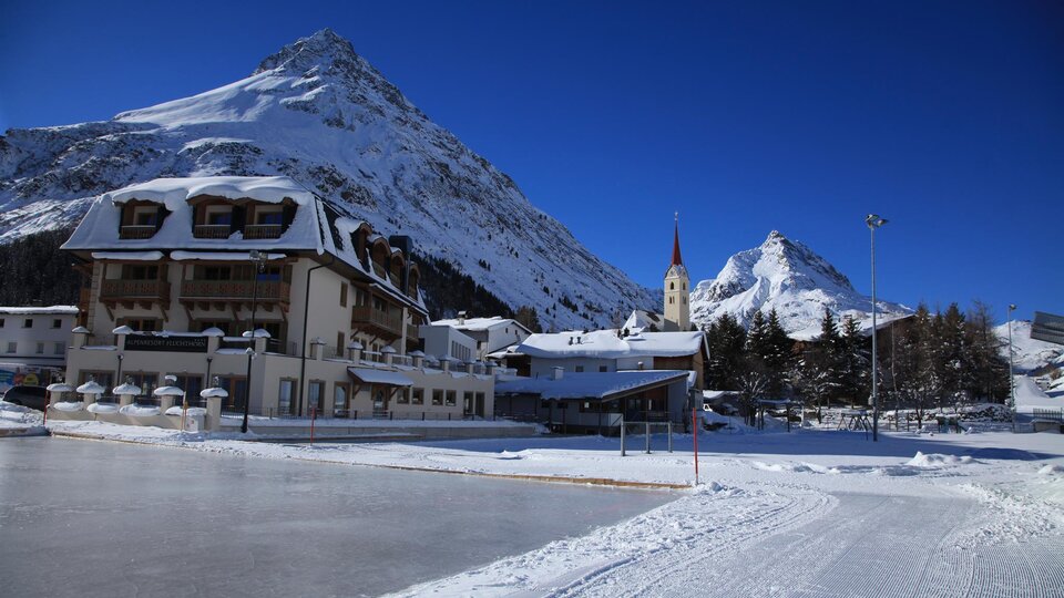 A picturesque mountain village in the snow with majestic mountains in the background. The sun is shining brightly in the blue sky. | © TVB Paznaun -  Ischgl