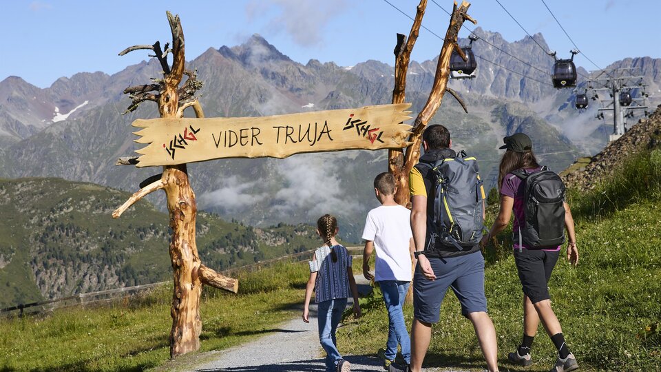 A hiking group is walking along a picturesque path to a sign that reads "Vider Truja". In the background, majestic mountains and a cable car can be seen.