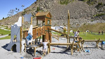 A wooden playground with slides and climbing elements.  
Children play and explore the surroundings on a beautiful, sunny day.