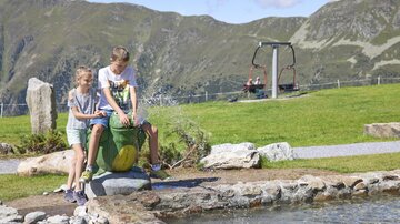 Two children are playing in a recreational area with water and stones. In the background, mountains and a chairlift can be seen.