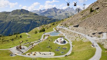 A picturesque mountain landscape with a modern cable car. In the foreground, there is a play area on green grass.