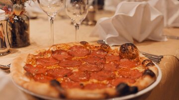 A deliciously topped pizza with salami on an elegant table. In the background, glasses and napkins are ready.