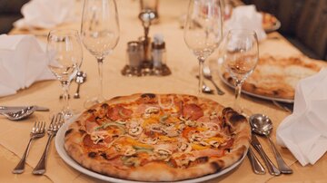 A table with elegant dishes and glasses. In the foreground is a large pizza with various toppings.