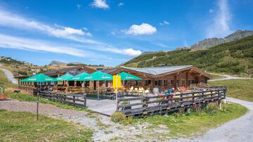 A cozy mountain cabin with a terrace and sun umbrellas. Surrounded by green meadows and mountains under a blue sky.