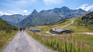 A picturesque path leads through a green landscape with mountains in the background. Hikers are walking on the trail to the next hut.