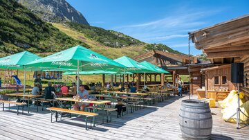 A beautiful outdoor terrace with tables and green sun umbrellas in the mountains. The surroundings are green and picturesque, ideal for a relaxed meal outside.