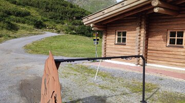 A rustic wooden house in a mountainous landscape. In the foreground stands a metal sign marking the entrance.