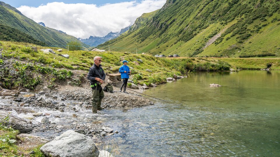 Two fishermen stand on the shore of a clear body of water and fish. The surroundings are green and inviting.