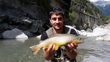 A fisherman stands on the bank of a river, proudly holding a large fish in his hands. In the background, rocks and trees can be seen.