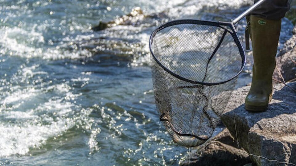 A fisherman stands on the shore of a body of water and holds a net in hand. The water flows sparkling in the background.