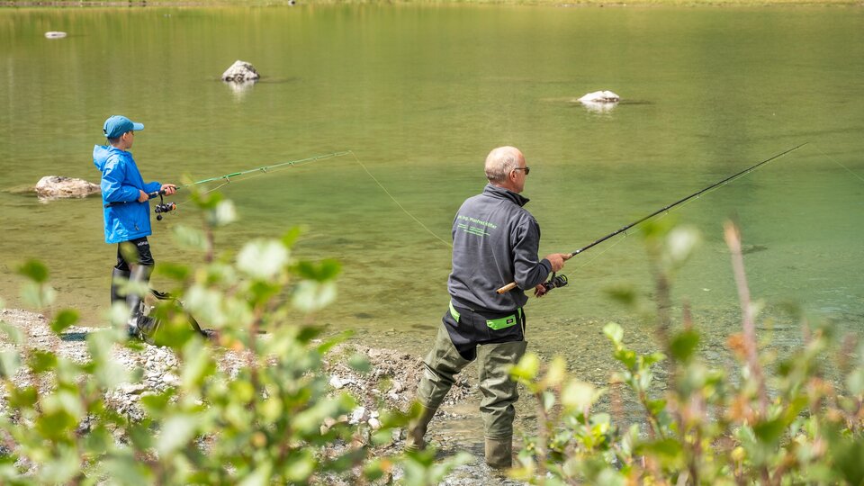 Two anglers are standing by the shore of a clear body of water. In the background, green meadows and stones can be seen.