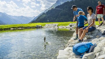 A boy is fishing with his father at a clear pond in the mountains. In the background, green meadows and tall mountains are visible.