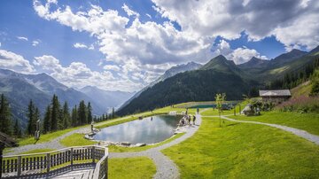 A picturesque landscape with a clear pond and green meadows. In the background, majestic mountains and a blue sky with white clouds can be seen.
