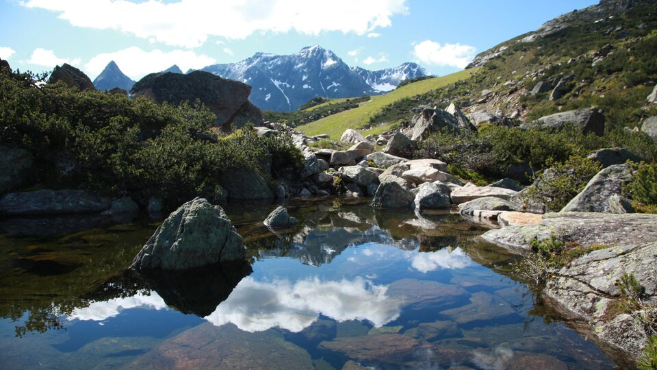 Eine malerische Berglandschaft mit hohen Gipfeln und klarem blauen Himmel. Im Vordergrund spiegelt sich das Gelände im ruhigen Wasser eines kleinen Teichs.