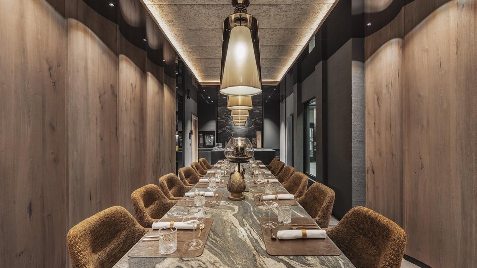 An elegant dining area with a long table and cozy chairs. The atmosphere is modern and stylish, illuminated by large lamps.