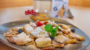 A plate with sweet pancakes, dusted with powdered sugar and garnished with berries and mint. There is also a bowl of fruit sauce.
