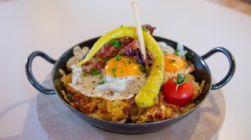 A hearty dish in a black pan with a fried egg, crispy bacon, and potatoes. Garnished with a bell pepper and a cherry tomato.