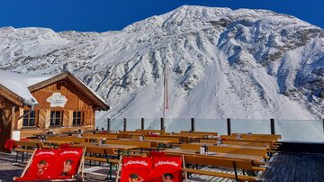 A mountain hut with a wooden terrace and several tables and chairs stands against an impressive mountain backdrop. The snow covers the landscape under a clear blue sky.