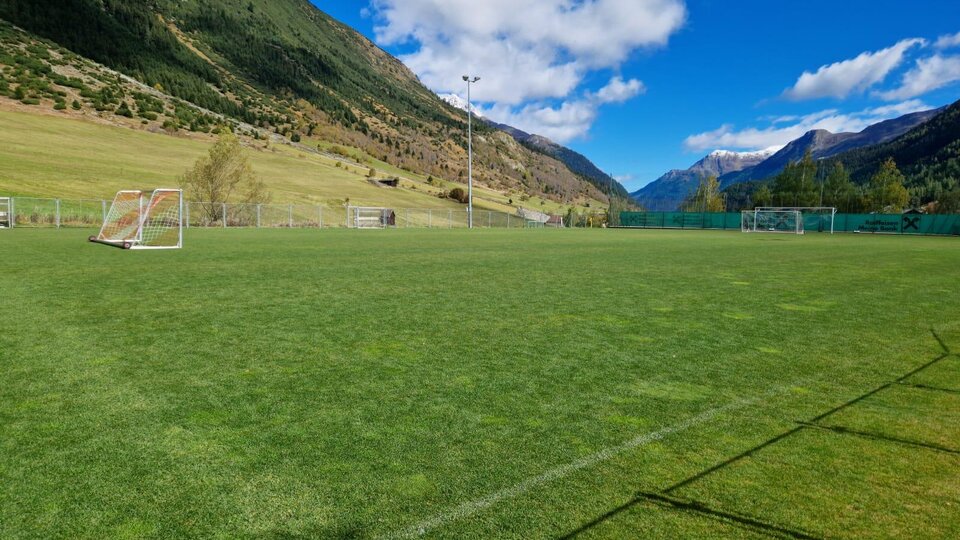 A green football field in a picturesque mountain landscape. In the background, high mountains and a clear blue sky can be seen.