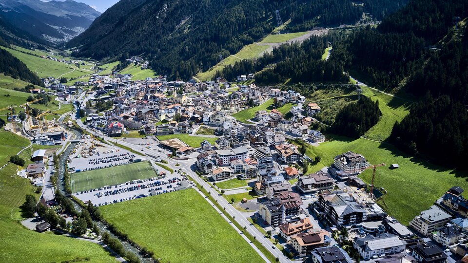 A picturesque alpine town in a green valley, surrounded by mountains. The buildings are closely packed, and there are many parking spaces and streets.